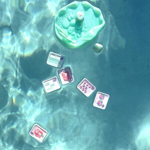 May include: A light green, circular, textured floating tray with a small die, accompanied by several smaller square white trays containing various colorful beads, charms, and small game pieces, all floating on the surface of clear blue water.