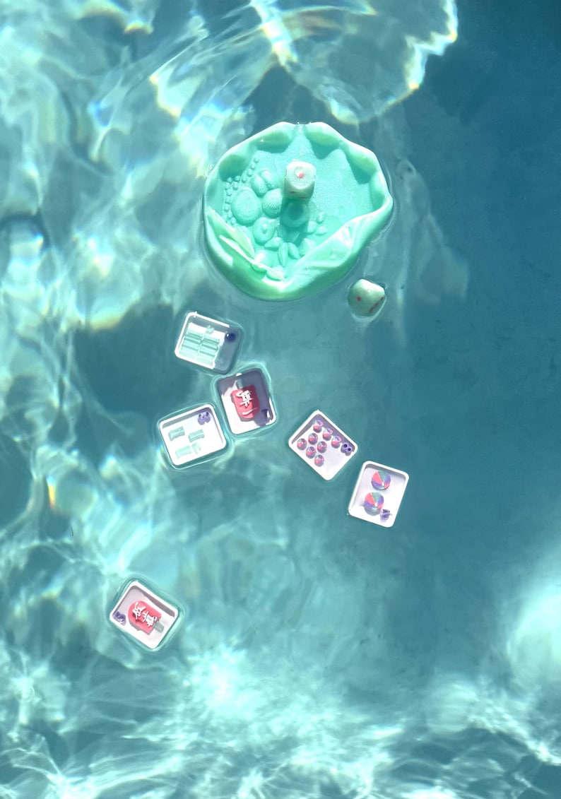 May include: A light green, circular, textured floating tray with a small die, accompanied by several smaller square white trays containing various colorful beads, charms, and small game pieces, all floating on the surface of clear blue water.
