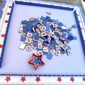 May include: A game board with a light purple surface and a blue scalloped border featuring red stars. A pile of square tiles with blue backs and white fronts displaying red, white, and blue graphics sits in the center with a red star-shaped dice cup.