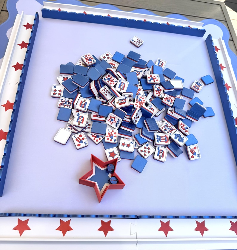 May include: A game board with a light purple surface and a blue scalloped border featuring red stars. A pile of square tiles with blue backs and white fronts displaying red, white, and blue graphics sits in the center with a red star-shaped dice cup.