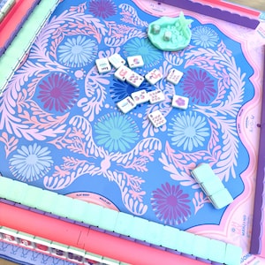 May include: A Mahjong game set featuring a blue play mat with a pink and purple floral pattern. The set includes light green tile racks, a light green dice cup, and white Mahjong tiles with various colorful symbols and text markings.