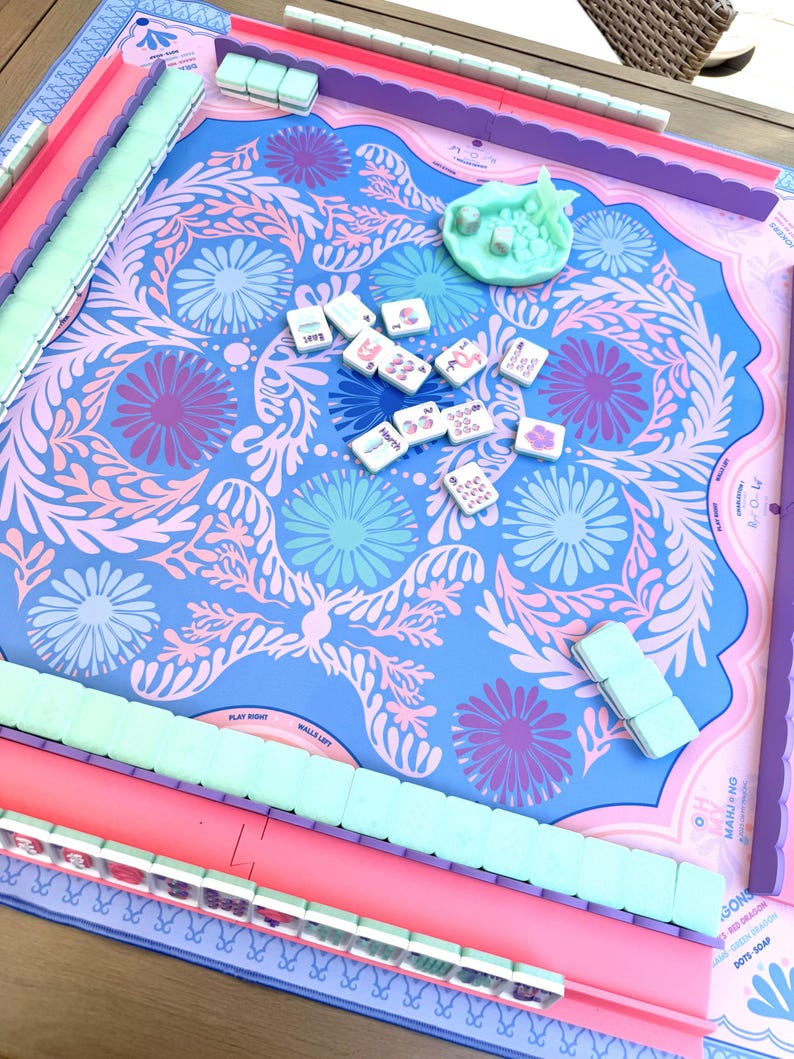 May include: A Mahjong game set featuring a blue play mat with a pink and purple floral pattern. The set includes light green tile racks, a light green dice cup, and white Mahjong tiles with various colorful symbols and text markings.