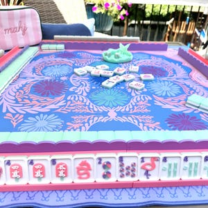 May include: A Mahjong game set featuring a blue and pink floral patterned play mat with scalloped edges. The set includes white tiles with pink and blue graphics, a light blue dice cup, and a pink and white striped zippered storage pouch with the text mahj.