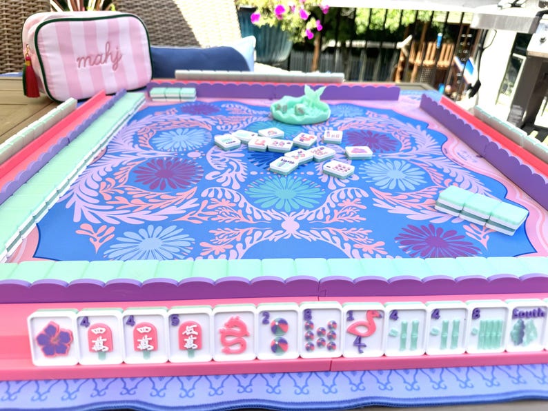 May include: A Mahjong game set featuring a blue and pink floral patterned play mat with scalloped edges. The set includes white tiles with pink and blue graphics, a light blue dice cup, and a pink and white striped zippered storage pouch with the text mahj.