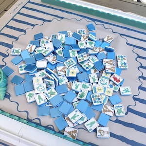 May include: A set of square Mahjong tiles with blue backs and white faces featuring colorful illustrations of sea creatures, plants, and sandcastles, scattered on a light-colored play mat with blue stripes and the text PLAY RIGHT printed on it.