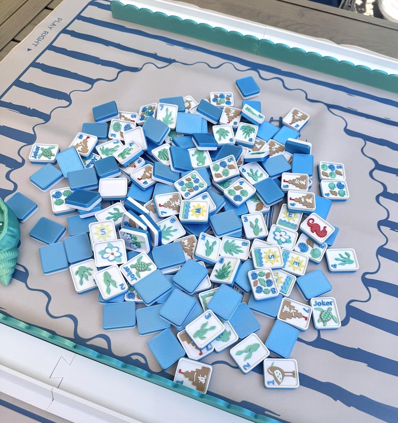 May include: A set of square Mahjong tiles with blue backs and white faces featuring colorful illustrations of sea creatures, plants, and sandcastles, scattered on a light-colored play mat with blue stripes and the text PLAY RIGHT printed on it.