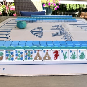 May include: A Mahjong set featuring white tiles with colorful beach-themed illustrations including seashells, seahorses, and sandcastles, arranged on a white play mat with blue striped borders and a central graphic of a beach umbrella and chairs.