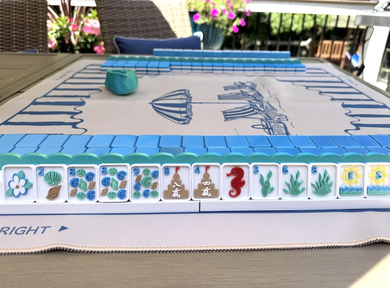 May include: A Mahjong set featuring white tiles with colorful beach-themed illustrations including seashells, seahorses, and sandcastles, arranged on a white play mat with blue striped borders and a central graphic of a beach umbrella and chairs.