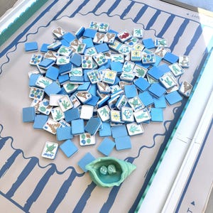 May include: A mahjong game set featuring a light gray play mat with blue stripes and directional text, a collection of blue and white tiles with sea-themed illustrations, and a light green seashell-shaped dice cup containing two dice.