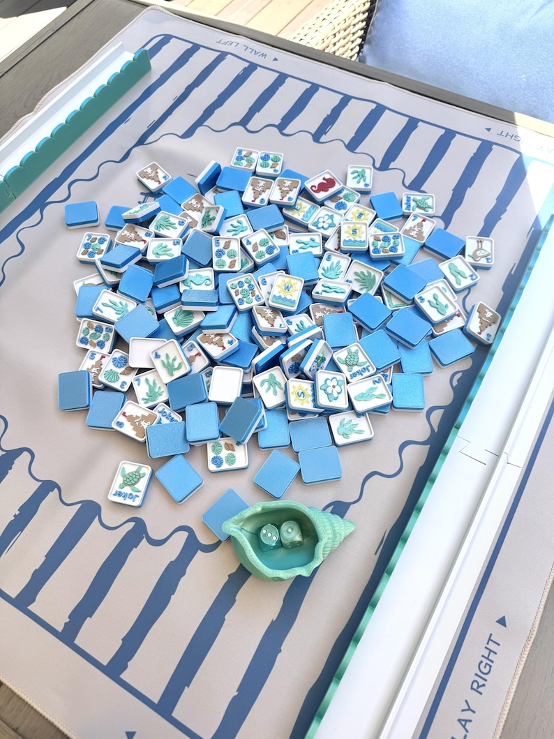 May include: A mahjong game set featuring a light gray play mat with blue stripes and directional text, a collection of blue and white tiles with sea-themed illustrations, and a light green seashell-shaped dice cup containing two dice.