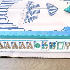May include: A Mahjong set featuring white tiles with colorful beach-themed illustrations, including seashells, starfish, and sandcastles, arranged on a white play mat with blue striped borders and a central beach umbrella and chair graphic.