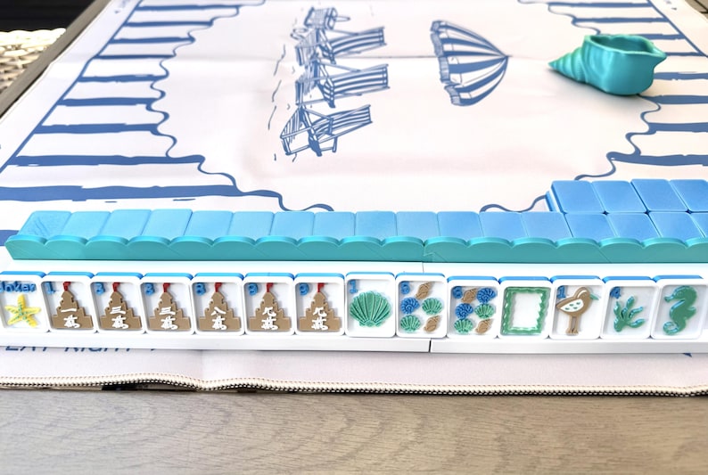May include: A Mahjong set featuring white tiles with colorful beach-themed illustrations, including seashells, starfish, and sandcastles, arranged on a white play mat with blue striped borders and a central beach umbrella and chair graphic.