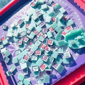 May include: A set of light mint green mahjong tiles scattered across a purple game board with scalloped edges. The tiles feature various red and blue symbols, characters, and numbers. A matching mint green tile holder sits on the right side.