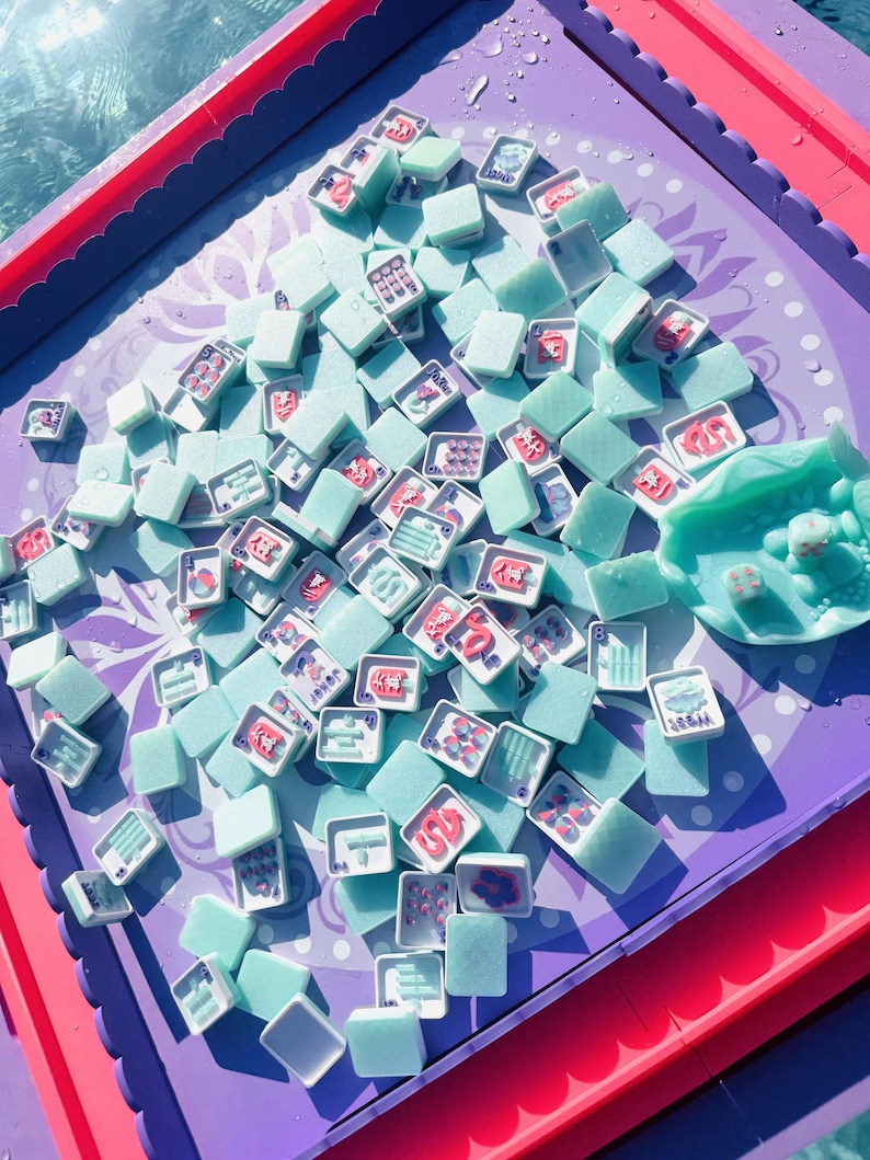 May include: A set of light mint green mahjong tiles scattered across a purple game board with scalloped edges. The tiles feature various red and blue symbols, characters, and numbers. A matching mint green tile holder sits on the right side.