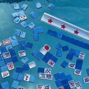 May include: A collection of blue square tiles and white rectangular tiles with red star and rocket graphics floating in water. A long white rack with red star accents is positioned near the top, featuring a scalloped blue edge along the side.
