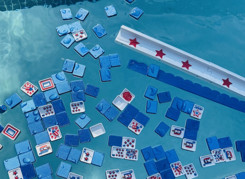 May include: A collection of blue square tiles and white rectangular tiles with red star and rocket graphics floating in water. A long white rack with red star accents is positioned near the top, featuring a scalloped blue edge along the side.