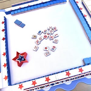 May include: A square game board with a white surface and a blue scalloped border featuring red stars. The board includes blue tile racks, a red star-shaped dice cup, and scattered game tiles decorated with patriotic red, white, and blue graphics.