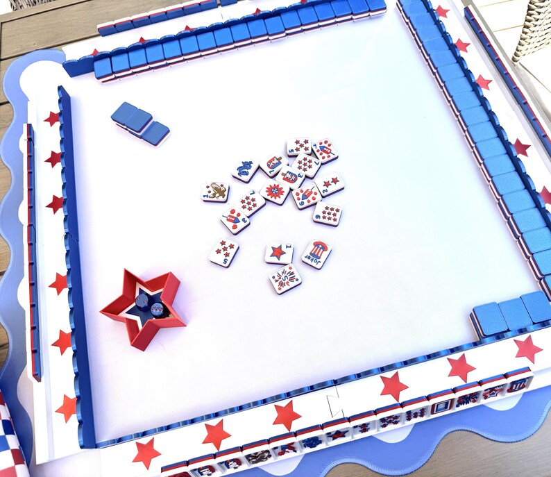 May include: A square game board with a white surface and a blue scalloped border featuring red stars. The board includes blue tile racks, a red star-shaped dice cup, and scattered game tiles decorated with patriotic red, white, and blue graphics.