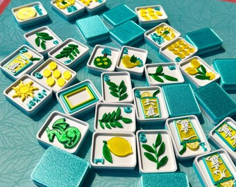 May include: A collection of mahjong tiles featuring white faces with colorful embossed designs including lemons, leaves, a dragon, a sun, and various symbols, paired with metallic teal backs, scattered across a matching teal and pink patterned mat.