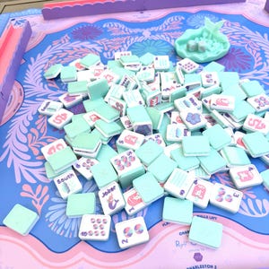 May include: A Mahjong game set featuring light mint green tiles with colorful illustrations and text, scattered across a blue and pink patterned game mat. The set includes a matching mint green dice tray with two dice and pink tile racks.