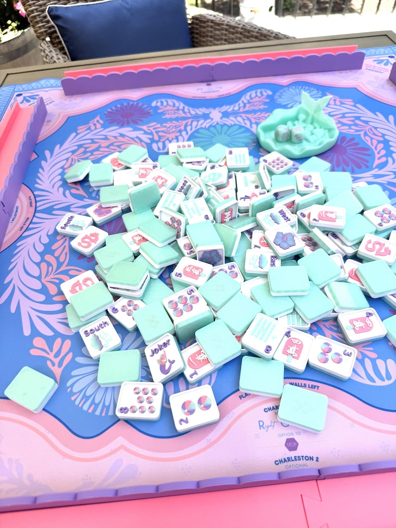 May include: A Mahjong game set featuring light mint green tiles with colorful illustrations and text, scattered across a blue and pink patterned game mat. The set includes a matching mint green dice tray with two dice and pink tile racks.