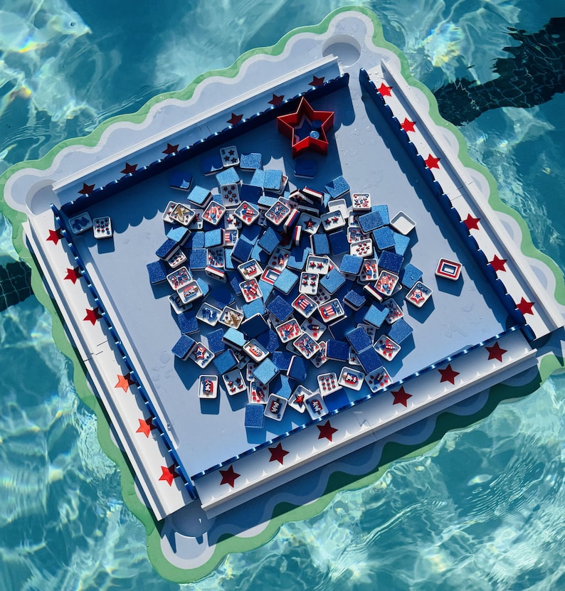 May include: A square floating game board with a light blue surface and a scalloped green border. The board features a red star-shaped container and numerous small blue tiles with patriotic red, white, and blue graphic designs scattered across.