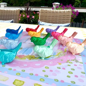 May include: A collection of twelve small, three-dimensional bird figurines in various metallic and solid colors, including blue, green, yellow, red, purple, pink, and peach, arranged in a circular pattern on a light-colored surface.