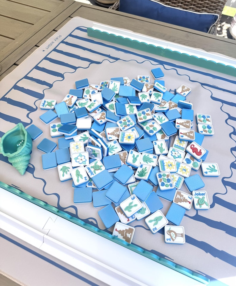 May include: A Mahjong game set featuring a light gray mat with blue wave patterns and the text PLAY RIGHT, a teal seashell dice cup, and a pile of square tiles with blue backs and white faces displaying various illustrations like turtles and flowers.