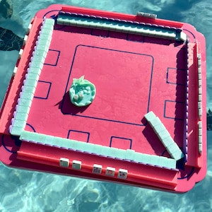 May include: A square, bright pink floating game board with a raised border and a central playing area. Pale green tiles are arranged along the edges and in a small pile in the center. The board features dark blue outlines and is wet from water.