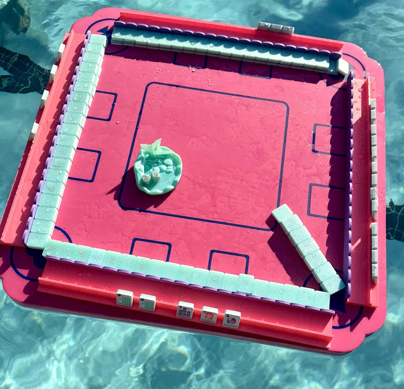 May include: A square, bright pink floating game board with a raised border and a central playing area. Pale green tiles are arranged along the edges and in a small pile in the center. The board features dark blue outlines and is wet from water.
