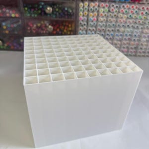 May include: A white plastic desk organizer featuring a grid of square compartments designed for storing pens, markers, or pencils. The structure is cubic with a solid exterior and an open top, providing multiple individual slots for upright storage.