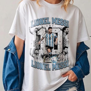 May include: A white t-shirt featuring a graphic print of Lionel Messi in an Argentina soccer jersey. The design includes the name Lionel Messi in light blue block letters, the number 10, and multiple illustrations of the athlete in action.