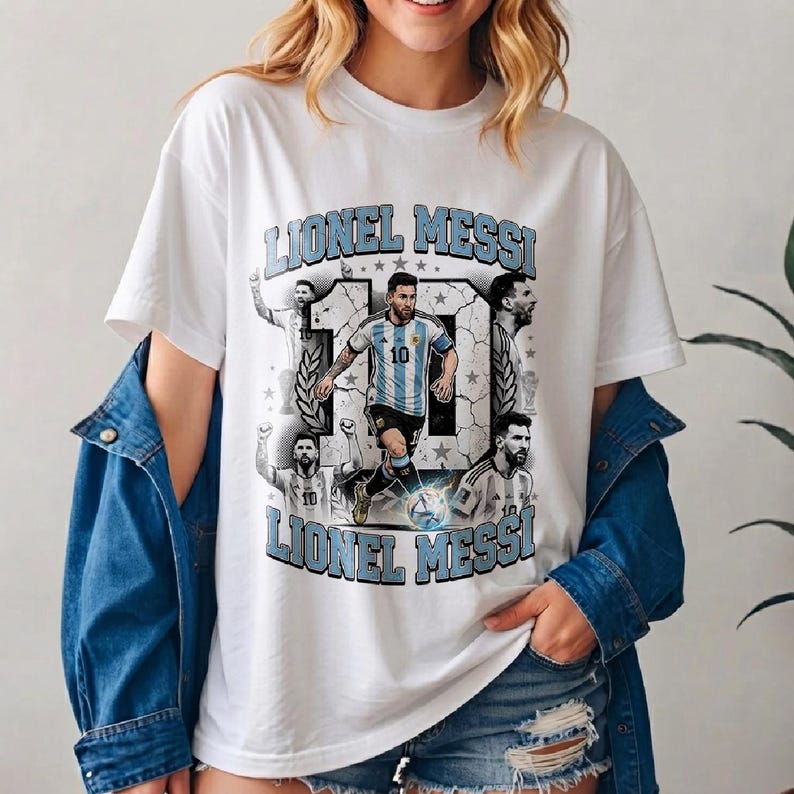 May include: A white t-shirt featuring a graphic print of Lionel Messi in an Argentina soccer jersey. The design includes the name Lionel Messi in light blue block letters, the number 10, and multiple illustrations of the athlete in action.