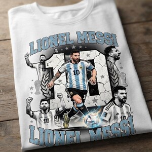 K&ouml;nnte beinhalten: Ein wei&szlig;es T-Shirt mit einem Grafikdruck von Lionel Messi im argentinischen Fu&szlig;balltrikot. Das Design enth&auml;lt den Text LIONEL MESSI, die Nummer 10, mehrere Bilder des Spielers in verschiedenen Posen und einen Fu&szlig;ball mit blauen Lichteffekten.