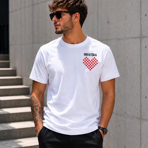 May include: A white short-sleeved t-shirt featuring a small graphic on the upper left chest. The graphic consists of the word "HRVATSKA" in black capital letters above a heart shape composed of a red and white checkered pattern.