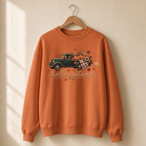 Könnte beinhalten: Ein orangefarbenes Sweatshirt mit Rundhalsausschnitt und einer Grafik eines dunkelgrünen Oldtimer-Pickups, der Kürbisse, Sonnenblumen und Herbstblätter geladen hat. Der Schriftzug "hello autumn" steht in Schreibschrift unter dem LKW.