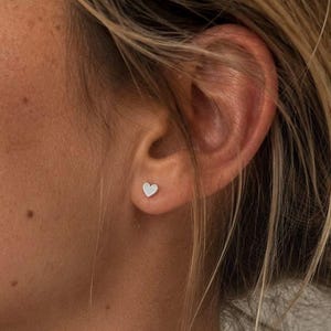 Könnte beinhalten: Ein kleiner, silberfarbener Ohrstecker in Herzform, der im Ohrläppchen getragen wird. Der Ohrstecker hat eine schlichte, glatte metallische Oberfläche und eine klassische Herzsilhouette. Der Text SM ESSENCE ist unten im Bild in Weiß zu sehen.