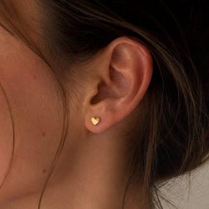 May include: A small, gold-coloured heart-shaped stud earring worn in the earlobe. The earring features a smooth, polished surface and a simple, minimalist design. The text SM ESSENCE is printed in white at the bottom of the image.