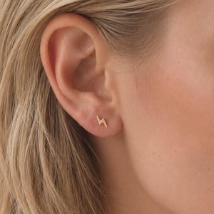 Peut inclure: Une petite boucle d'oreille à clou en or en forme d'éclair portée au lobe de l'oreille. La boucle d'oreille présente une finition métallique polie et un design angulaire simple. Le texte SM ESSENCE est imprimé en blanc au bas de l'image.