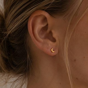Könnte beinhalten: Ein kleiner goldener Ohrstecker in Form eines Halbmondes, der im Ohrläppchen getragen wird. Der Ohrstecker hat eine glatte, polierte Oberfläche und eine geschwungene Form. Der Text SM ESSENCE ist unten im Bild zu sehen.
