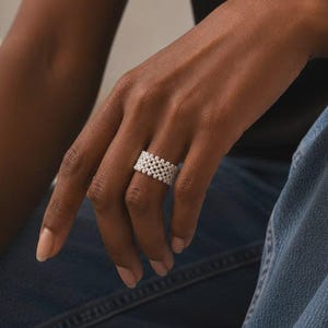 May include: A wide ring crafted from small, white, spherical beads arranged in a repeating, interconnected grid pattern. The band features a uniform, textured surface created by the dense placement of the beads. The text SM ESSENCE is visible.