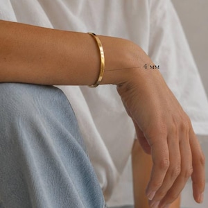 Può includere: Un braccialetto minimalista dorato, largo 4 mm, è indossato su un braccio. La persona indossa una maglietta bianca e jeans blu. Il nome del marchio "SM ESSENCE" è visibile in basso nell'immagine.