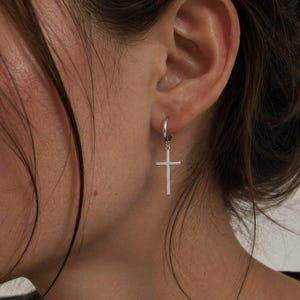 Peut inclure: Une boucle d'oreille créole argentée avec une breloque en forme de croix. Le bijou se compose d'un petit anneau circulaire poli qui s'attache au lobe de l'oreille, avec un pendentif en forme de croix fine et verticale suspendu à l'anneau.