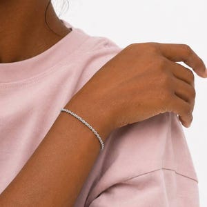 May include: A thin, silver-toned rope chain bracelet worn on the wrist. The bracelet features a twisted, textured metal link design. The text SM ESSENCE is printed in black at the bottom of the frame against a light pink t-shirt background.