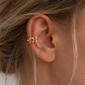 Pode incluir: Um ear cuff dourado com um design delicado de ramo, adornado com pequenas pedras transparentes de corte marquise. A peça está posicionada na cartilagem da orelha. O texto SM ESSENCE está impresso em branco na parte inferior da imagem.