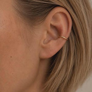 May include: A simple, smooth, gold-toned metal ear cuff worn on the cartilage of the ear. The piece features a minimalist, circular band design with a polished finish. The text SM ESSENCE is visible at the bottom of the image.