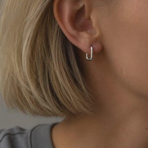 May include: A small, silver-toned rectangular hoop earring with a smooth, polished finish. The earring features a minimalist, geometric shape that secures through the earlobe. The text SM ESSENCE is printed in white at the bottom of the image.