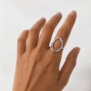 May include: A silver-toned ring featuring an open, irregular oval-shaped design with a smooth, polished finish. The ring is worn on the finger, showcasing its continuous, rounded metal band that forms a distinct, non-symmetrical loop shape.
