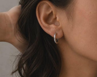 May include: A small, polished silver hoop earring worn in the earlobe. The earring features a smooth, circular design with a reflective metallic finish. The text SM ESSENCE is printed in white at the bottom centre of the image frame.