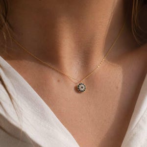 May include: A delicate gold chain necklace featuring a small, circular pendant. The pendant is encrusted with tiny, dark blue stones arranged in a circular pattern around a central dark stone, creating a detailed, eye-like motif on the jewellery piece.